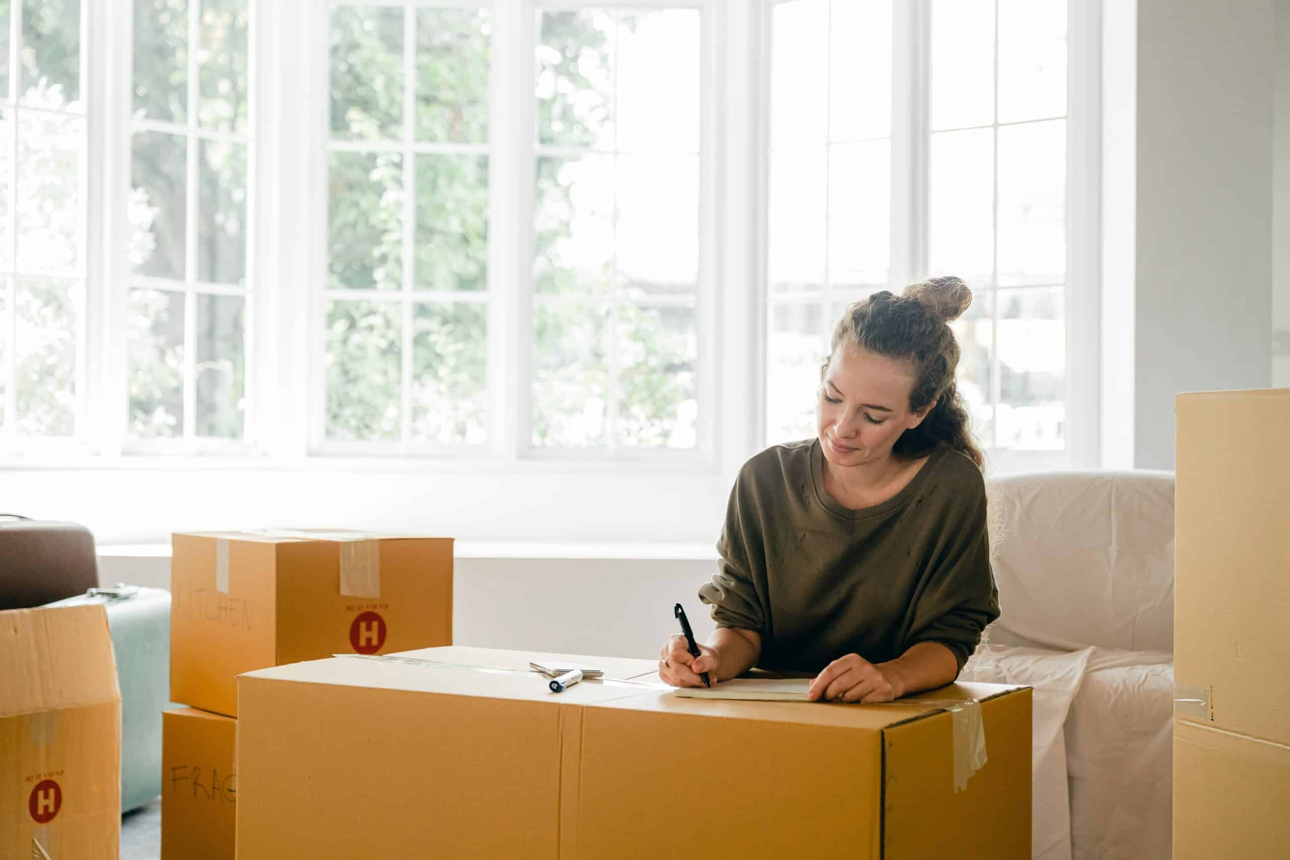 Woman writing on notepad surrounded by moving boxes.
